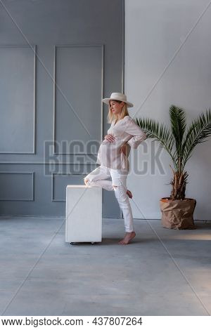 Fashion Pregnant Young Woman In White Hat Leaning Her Leg On A White Cube In A Gray, Stylish Studio