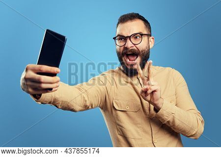 Excited Bearded Man Wearing Eyeglasses And Beige Shirt, Screaming Loud And Showing V Sign While Taki