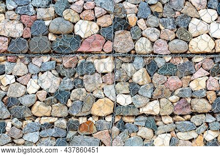 Stone Wall With Metal Grid And Rough Stones As Background. Rock Texture In Metal Grid. Gabion Net Wi
