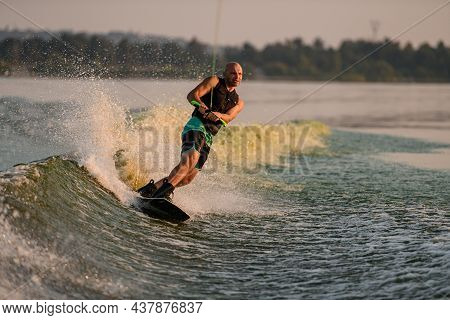Athletic Muscular Man Holds Rope And Energetically Riding Wakeboard On Splashing River Water.