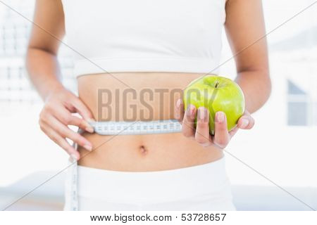 Close up mid section of a young woman measuring waist as she holds apple in fitness studio