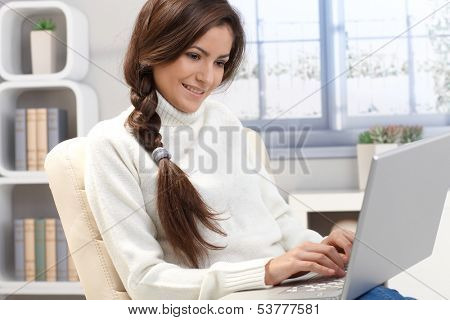 Portrait of long hair beauty typing on laptop computer at home, smiling, sitting in armchair in living room.