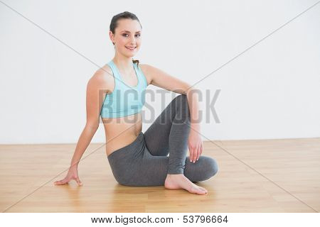 Full length portrait of a fit young woman sitting in cowface posture against wall in fitness studio
