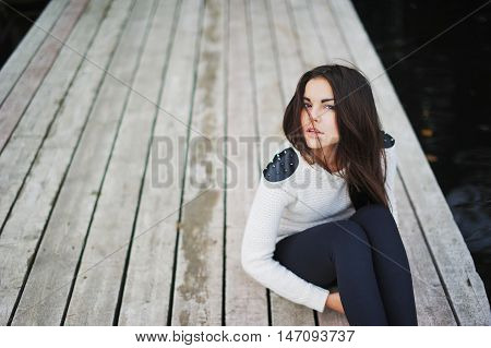 Portrait of a confident, beautiful brown-eyed brunette with long hair tousled by the wind, in a white jacket with spikes on his shoulders, sitting on the wooden walkways.