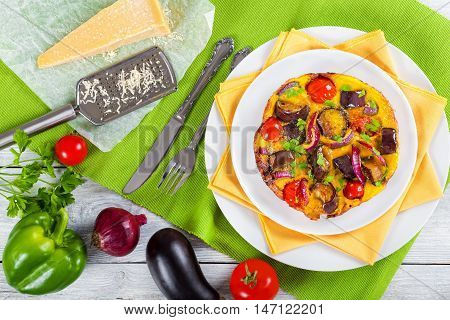 frittata with red onion eggplant tomato sprinkled with parsley on white dish with table napkin parmesan cheese and gratter on parchment paper view from above