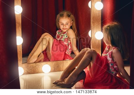 Cute little fashionista. Happy child girl try on outfits and mom's shoes looking at mirror.