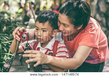 Portrait of young asian mother teaching her son photographing outdoors using digital camera. Happy family spending time together. Positive human emotion. Vintage effect and cream tone.