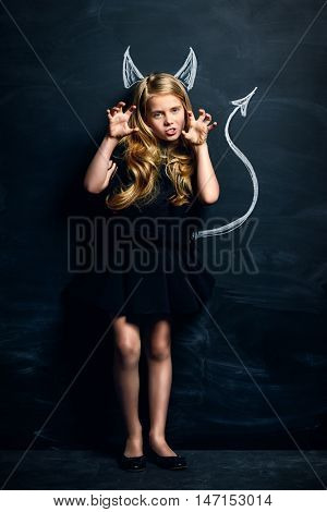 Funny little imp girl. Cute child girl posing with imp horns and tail drawn on a blackboard.