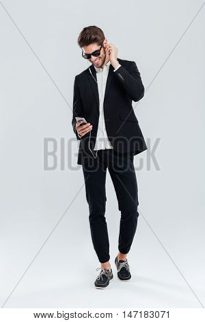 Full length portrait of a smiling happy young businessman litening music with earphones and smartphone over gray background