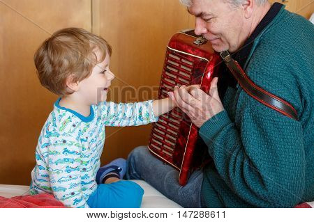 Happy blond little kid boy and his grandfather playing together with accordion. Senior man teaching his grandson, cute toddler to play with music instrument at home.
