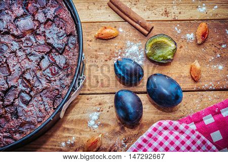 Chocolate clafoutis with plums in a baking pan, ripe fresh  plums and it`s stones, cinnamon, flour and cocoa powder on wooden background