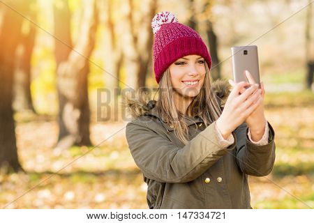 Beautiful young woman in jacket and knitted hat taking a selfie in autumn on smart phone