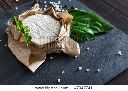 Cheese delikatessen closeup on black stone surface and wood. Camembert or brie circle in brown kraft paper decorated with basil