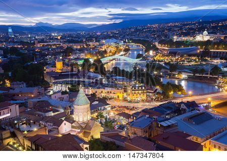 Scenic Top View Of Tbilisi, Georgia In Evening Lights Illumination. Metekhi Bridge, Surb Gevorg Church And All Famous Landmarks, Sightseeings Under Beautiful Blue Cloudy Sky.