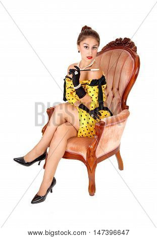 A lovely young woman sitting in a yellow dress in a pink armchair holding a long cigarette holder isolated for white background.