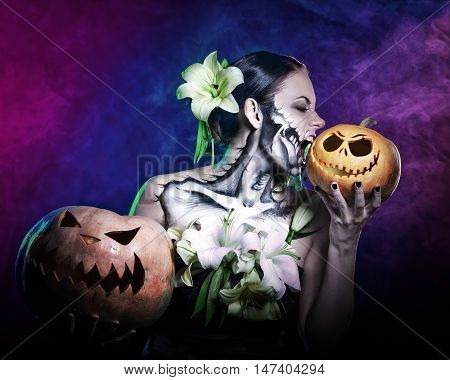 Young attractive girl with creative make-up for Halloween. Portrait close-up pumpkins. She bites pumpkin. Mysterious and frightening image of lilies. Witchcraft. Horrible. Jack-o'-lantern