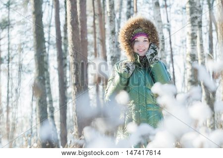 Cheerful young woman is holding woolly mittens near her head in winter forest outdoors