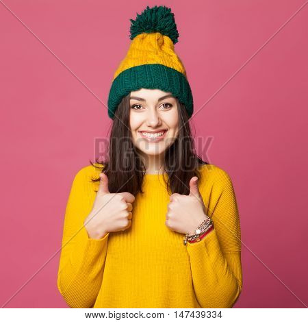 Happy smiling young girl in bright warm sweater and hat showing ok sign against a pink colorful wall. Autumn stylish clothes.