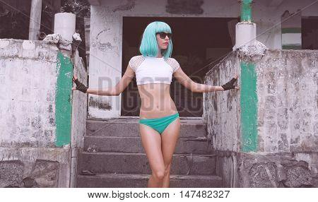 Sexy beautiful woman in modern futuristic style posing over abandoned building. Creative look of woman on the beach wearing bikini, black leather fingerless and blue wig
