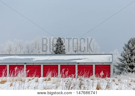 Multiple storage units with red doors next to woods in the winter.