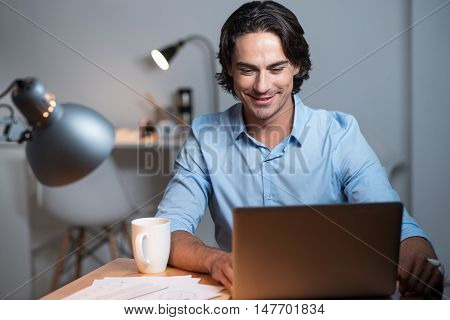 Dream work. Handsome positive young man sitting at the table and putting his lags on the table while using tablet.