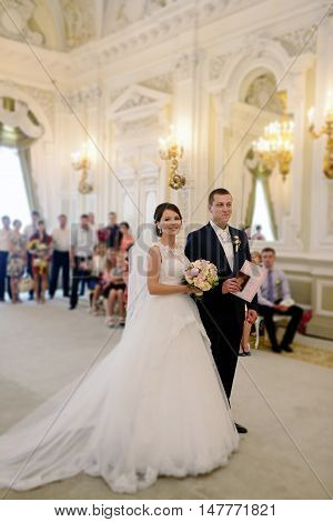Beauty bride and handsome groom are registering the marriage. Wedding couple is marrying. Beautiful model girl in white dress and in lace veil. Man in suit. Female and male portrait. Cute lady and guy