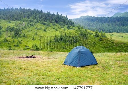 View of tent on meadow in mountains. Camping background. Camping and tent. Tourist tent on meadow green pine forest mountains and sky on the background. Adventure travel active lifestyle freedom outdoors.
