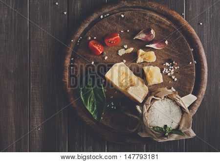 Cheese delikatessen closeup on rustic wood. Wooden desk with parmesan, camembert and brie cuts decorated with garlic, tomato and basil, top view image with copy space