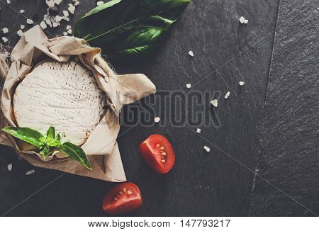 Cheese delikatessen closeup on black stone desk surface. Camembert or brie circle in brown kraft paper decorated with basil and pieces of cherry tomatoes, top view image with copy space