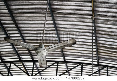 Old rusty gray metal ceiling fan hang on warehouse ceiling which covered with insulator to reduce temperature.