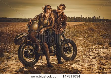 Young, stylish cafe racer couple on the vintage custom motorcycles in a field.