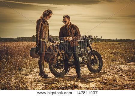 Young, stylish cafe racer couple on the vintage custom motorcycles in a field.