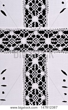 Latin cross motiv in whitework embroidery linen tablecloth. Reticella handicraft decoration with yarn. White stitching on white linen. Macro photo close up from above on black background.