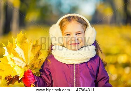 Happy little girl in earflaps with autumn leaves in the park
