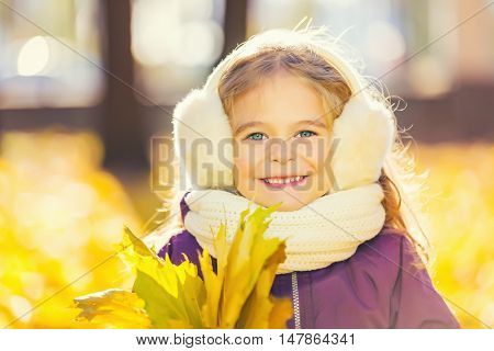 Happy little girl in earflaps with autumn leaves in the park