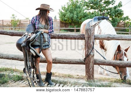 Happy beautiful young woman cowgirl in hat standing and holding saddle for riding horse