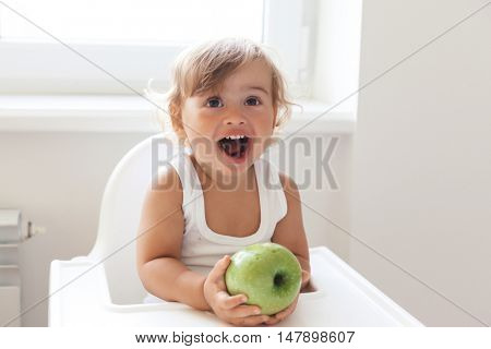 Cute baby 1,4 years old sitting on high children chair and eating fruit alone in white kitchen