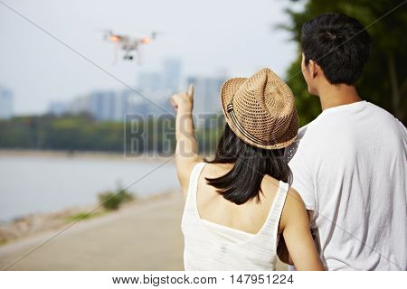 young asian woman pointing to a flying drone operated by her friend or partner