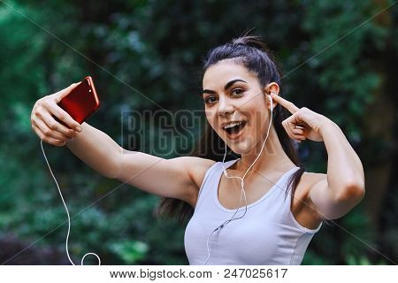 Young Girl Is Taking Picture Of Herself Using Red Cell Phone And Listening To The Music Using White 