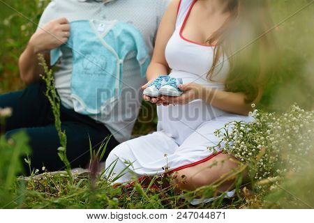 Young Couple With Pregnant Woman Looking Presents Children Clothes And Infant Shoes Together - Happy
