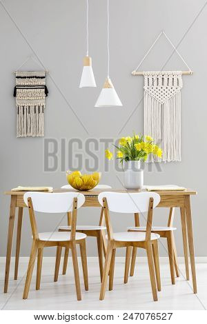 White Chairs At Wooden Table With Yellow Flowers In Grey Dining Room Interior With Lamps. Real Photo
