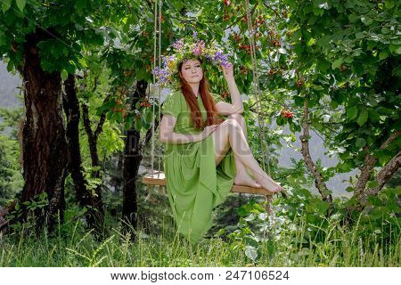Young, Beautiful Woman In A Green Dress With A Wreath Of Wild Flowers Sits On A Homemade Swing In A 