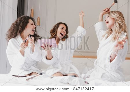 Young smiling women 20s wearing housecoat drinking champagne and applying makeup during bridal shower while resting in luxury bedroom at hotel room