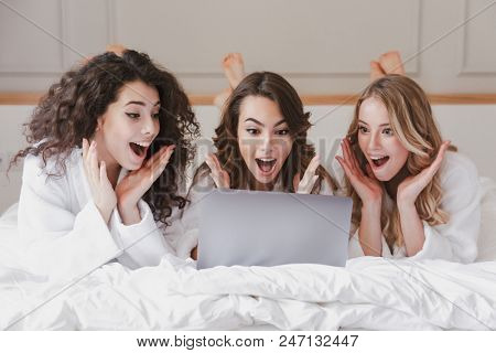Image of caucasian smiling women 20s wearing white housecoat lying in luxury bedroom at home and using silver laptop during bachelorette party