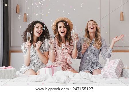 Portrait of beautiful excited women 20s wearing dresses celebrating bridal shower in posh apartment with champagne and falling confetti