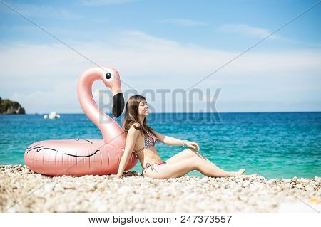 Young Woman With Bikini Sitting Close To Giant Inflated Flamingo Float On A Beach With Turquois Wate