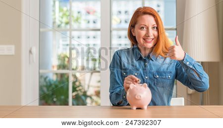 Redhead woman saves money in piggy bank at home happy with big smile doing ok sign, thumb up with fingers, excellent sign