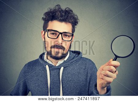 Bearded Scrupulous Man In Glasses Holding Magnifying Glass Looking At Camera On Gray Background.