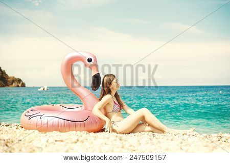 Young Woman With Bikini Sitting Close To Giant Inflated Flamingo Float On A Beach With Turquois Wate