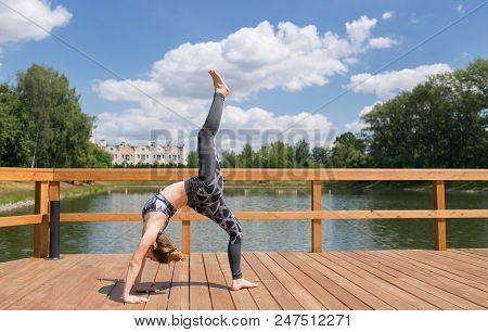 Young Slim Girl In Grey Sport Wear Doing Yoga On The Bridge. Flexible Young Girl With Red Hair.